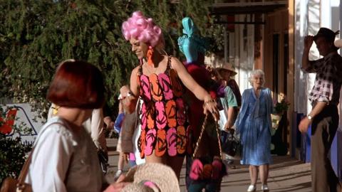 Hugo Weaving with a bright pink wig and wearing a dress made out of pink and orange flip-flops. He is walking down a footpath in drag make-up and attracting surprised looks from passersby