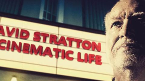 Close up of David Stratton standing next a sign that reads: David Stratton a cinematic life