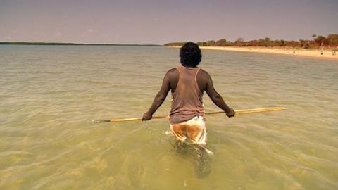 An Indigenous man walking through the water holding a spear