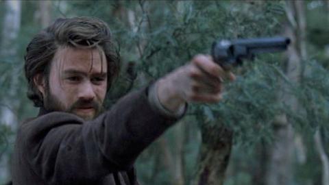 Heath Ledger as Ned Kelly holds a gun out in front of him.